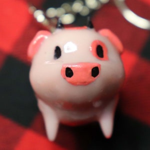 K&ouml;nnte beinhalten: Ein rosa Schweine-Schl&uuml;sselanh&auml;nger mit schwarzen Augen und einer rosa Nase. Das Schwein hat zwei schwarze H&ouml;rner auf dem Kopf und ist aus einem glatten, gl&auml;nzenden Material gefertigt.
