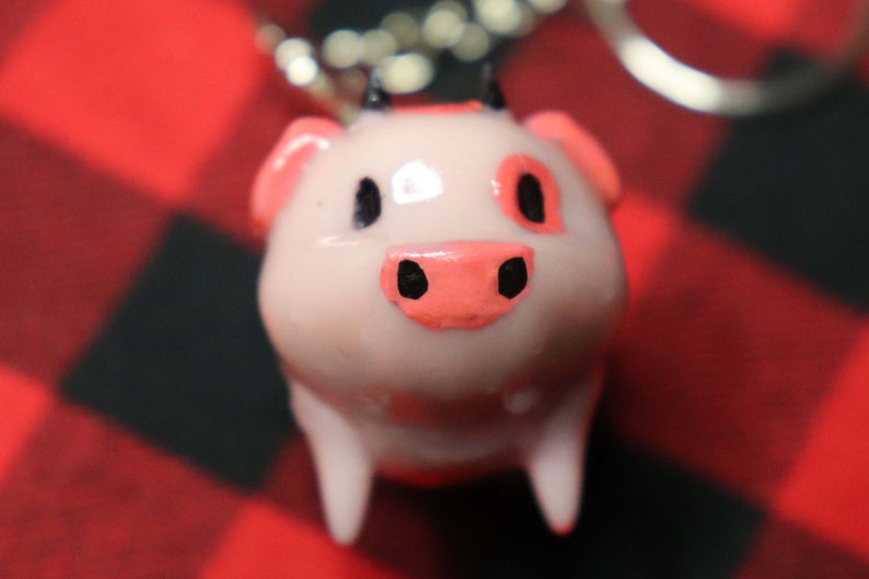 K&ouml;nnte beinhalten: Ein rosa Schweine-Schl&uuml;sselanh&auml;nger mit schwarzen Augen und einer rosa Nase. Das Schwein hat zwei schwarze H&ouml;rner auf dem Kopf und ist aus einem glatten, gl&auml;nzenden Material gefertigt.