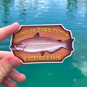 May include: A sticker featuring a catfish illustration with the text "Come On Down To The Catfish Cabin" in a decorative font. The catfish is depicted in shades of gray and purple against a brown background, with a water background.