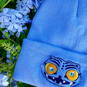 May include: A light blue beanie hat with a folded brim. The hat features an embroidered tiger face with yellow eyes and a dark blue outline. The hat is set against a backdrop of blue flowers and green foliage.
