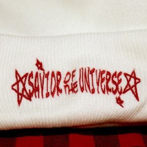May include: A white knit beanie featuring red embroidery on the folded cuff. The text reads SAVIOR OF THE UNIVERSE in a stylized font, flanked by two five-pointed star outlines. The embroidery has a textured appearance against the white fabric.