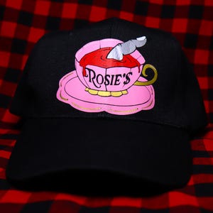 Puede incluir: Gorra de béisbol negra con un diseño de taza de té rosa, el texto "Rosie's" y un cuchillo plateado. La taza de té está llena de líquido rojo y se apoya en un platillo rosa. El fondo es un patrón de cuadros rojos y negros.
