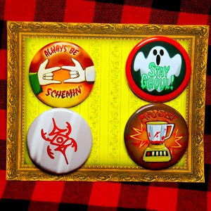 May include: Four circular pin badges with different designs and text, including "ALWAYS BE SCHEMIN", a ghost with "STAY GHOUL!", a red symbol, and a blender with "ARISE!". The badges are in a gold frame.