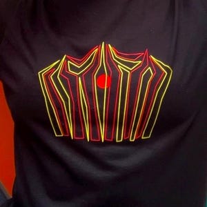 May include: Black t-shirt featuring a geometric design in yellow and red. The design has a central red circle and a series of lines. The shirt is a crew neck style.