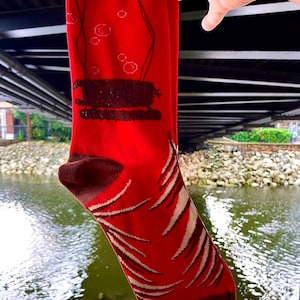 Puede incluir: Un calcetín rojo de caña media con un gráfico marrón oscuro de un submarino sumergido con burbujas ascendentes. El calcetín tiene un puño y un talón de color marrón oscuro. La parte inferior muestra patrones blancos de rayas diagonales.