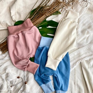 Könnte beinhalten: Drei Paar Babyhosen in verschiedenen Farben: rosa, blau und weiß. Die Hosen sind aus weichem, dehnbarem Stoff und haben eine bequeme Passform.