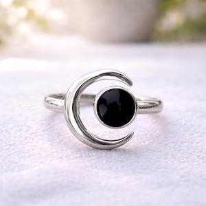 Può includere: Anello d'argento con un design a mezzaluna e una pietra nera circolare al centro. L'anello ha una finitura lucida ed è posto su uno sfondo bianco morbido.