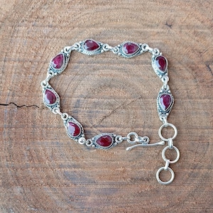 Op de afbeelding: Een zilveren armband met een rood edelsteen ontwerp. De armband heeft een karabijn sluiting.