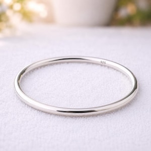 Könnte beinhalten: Ein poliertes silbernes Armreif-Armband. Das Armband ist ein einfaches, glattes Band mit rundem Profil. Der Text "925" ist auf der Oberfläche eingeprägt. Das Armband wird auf einer weißen Oberfläche präsentiert.