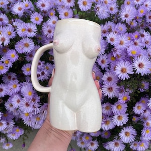 Puede incluir: Una taza de cerámica blanca con forma de torso humano. La taza tiene un asa y está decorada con un sutil acabado brillante.