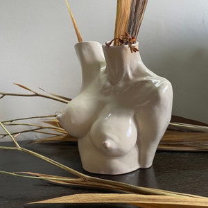 Könnte beinhalten: Eine weiße Keramikvase in Form eines Torsos mit getrockneten Blumen in der Öffnung. Die Vase hat eine glatte, glänzende Oberfläche.
