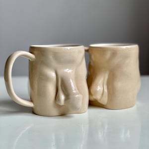 May include: Two ceramic mugs shaped like human torsos. The mugs are a light beige color and have a glossy finish.