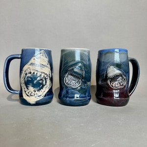 May include: Three ceramic mugs with a blue and white glaze. Each mug features a white shark with an open mouth. The mugs are all different sizes and shapes.