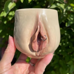 May include: A beige ceramic mug with a detailed vulva design, featuring pink and brown tones. A silver-colored ring is placed in the center of the design. The mug is held in a hand against a blurred green background.