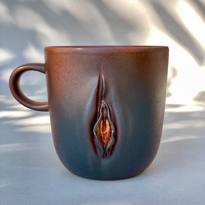 May include: A brown ceramic mug with a textured, abstract design. The design features a raised, textured, and slightly orange-colored shape in the center of the mug.