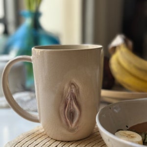 May include: A beige ceramic mug with a brown and pink abstract design. The mug has a handle and is sitting on a wooden surface.