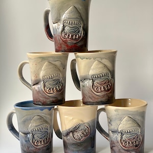 Pode incluir: Seis canecas de cerâmica com um design de tubarão. As canecas são em vários tons de azul, cinza e marrom. Cada caneca apresenta um tubarão branco com a boca aberta e dentes afiados. As canecas estão empilhadas em forma de pirâmide.