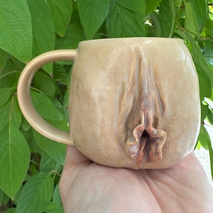 May include: A beige ceramic mug with a sculpted vulva design. The mug has a rounded shape with a large handle and a detailed, textured surface. The vulva is rendered in shades of brown and pink.