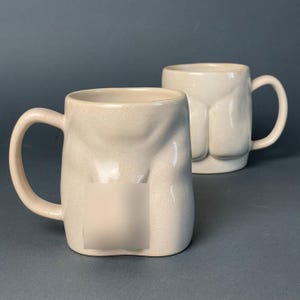 Puede incluir: Dos tazas de cerámica beige con un acabado texturizado y moteado. Cada taza presenta un diseño de torso anatómico esculpido en la parte frontal, con un asa curva en el lateral. Una de las tazas tiene una sección borrosa en el centro.
