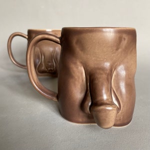 May include: Two brown ceramic mugs with a unique, sculpted design. The mugs have a handle and a rounded body.