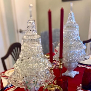 May include: A festive table setting featuring a red tablecloth with a blue and white patterned border. The table is set with clear glass Christmas tree-shaped centerpieces, red candles, and gold-rimmed china. Wine glasses and teacups complete the elegant holiday display.