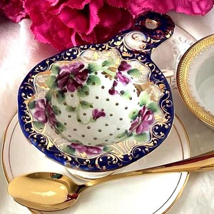 May include: A porcelain tea set with a floral tea strainer, a small bowl, a saucer, and a gold spoon. The tea strainer has a blue rim and is decorated with purple flowers. The bowl and saucer have gold trim. The spoon is gold.
