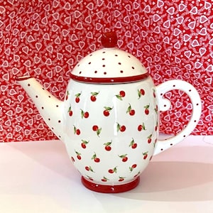 May include: White ceramic teapot with a red lid and red cherry pattern. The teapot has a red rim and a red base.