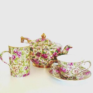 May include: A porcelain tea set featuring a teapot, a tall mug, and a teacup with a matching saucer. Each piece is decorated with a dense pattern of pink and yellow roses, green leaves, and gold trim on the rims, handles, and teapot lid.
