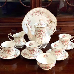 May include: A complete vintage tea set featuring a teapot, creamer, sugar bowl, serving plate, and six teacups with saucers. The set is white with a pink floral pattern and gold trim, perfect for a tea party.