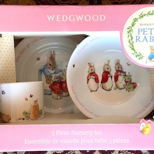 May include: A Wedgwood 3-piece nursery set in a pink box featuring Beatrix Potter Peter Rabbit illustrations. The set includes a white ceramic mug and two bowls decorated with colorful rabbit characters and text. The packaging displays the Peter Rabbit logo.