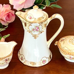 May include: A vintage porcelain tea set with a white base and gold trim, featuring floral accents. The set includes a creamer, a covered sugar bowl, and a tall coffee pot with a lid. Pink roses are in the background.