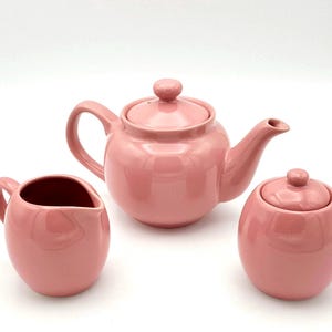 May include: A pink ceramic teapot, creamer, and sugar bowl set. The teapot has a round body and a spout. The creamer and sugar bowl are both round.