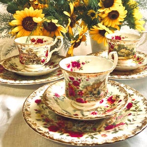 May include: Three sets of teacups and saucers with floral patterns. The cups and saucers are white with gold trim and feature a cottage scene with red and pink flowers. The cups have a handle and the saucers are round.