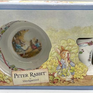May include: A boxed Peter Rabbit by Wedgwood baby dish set. The set includes a bowl and a cup, both decorated with illustrations of Peter Rabbit. The box has a light blue border and features the Peter Rabbit logo.