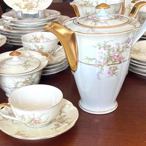 May include: A white porcelain tea set with gold trim and floral accents. The set includes a teapot, cups, saucers, plates, and serving dishes. The floral design features pink and purple flowers with green leaves.