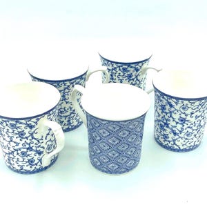 May include: Five white ceramic mugs with blue floral and geometric patterns. Each mug has a white handle and a white interior. The mugs are arranged on a white surface, showcasing their decorative designs.