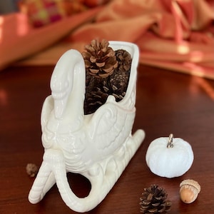 May include: White ceramic swan-shaped sleigh with pine cones inside. The sleigh is on a brown wooden surface with a white pumpkin and pine cones.