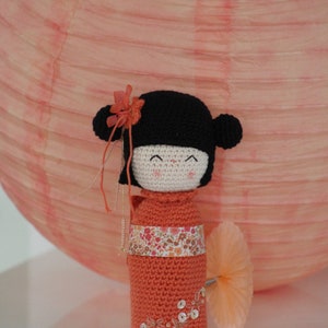 Op de afbeelding: Een gehaakte pop met zwart haar, een roze bloem en een perzikkleurige jurk met bloemenborduursel en pailletten. De pop staat voor een perzikkleurige papieren lantaarn.