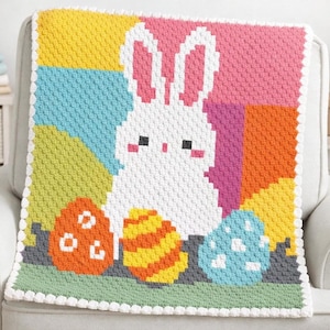 Puede incluir: Una colorida manta de Pascua tejida a ganchillo con un conejito blanco y tres huevos decorados. La manta tiene un borde blanco y está hecha con varios colores, incluyendo rosa, azul, naranja, amarillo y verde. El conejito tiene orejas y mejillas rosas.