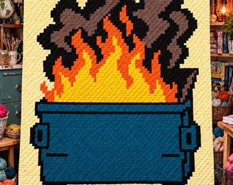May include: A rectangular crochet blanket featuring a pixelated design of a blue dumpster on fire. The flames are rendered in bright yellow and orange, with dark gray smoke rising above. The background of the blanket is a solid pale yellow.