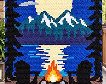 May include: A rectangular crochet blanket featuring a pixelated landscape scene. The design displays a large yellow moon, snow-capped mountains, a blue lake, and a bright orange campfire in the foreground, framed by tall black pine tree silhouettes.