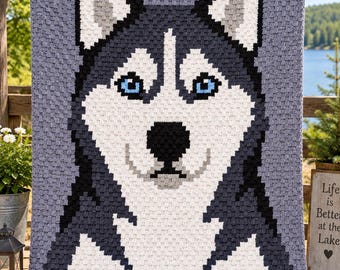 May include: A rectangular crochet blanket featuring a pixelated portrait of a Siberian Husky dog. The design uses shades of grey, black, and white yarn to depict the dog's face, with bright blue eyes and a black nose set against a light grey background.