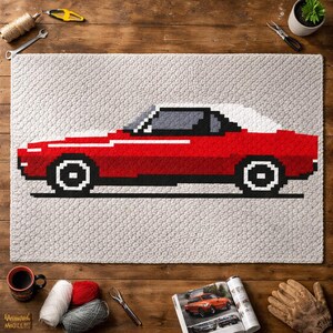 May include: A red pixelated car design on a light gray rug. The car has a white roof and black wheels. The rug is surrounded by craft supplies, including yarn, a coffee mug, and a magazine with a car image.