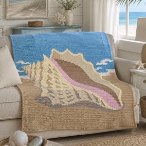 May include: A rectangular crochet blanket featuring a large conch shell design centered on a sandy beach background under a bright blue sky. The shell is rendered in shades of cream, brown, and pink, with a textured stitch pattern throughout.