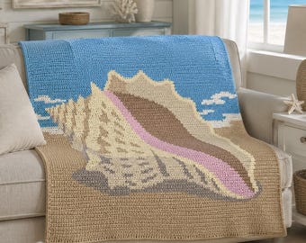 May include: A rectangular crochet blanket featuring a large conch shell design centered on a sandy beach background under a bright blue sky. The shell is rendered in shades of cream, brown, and pink, with a textured stitch pattern throughout.
