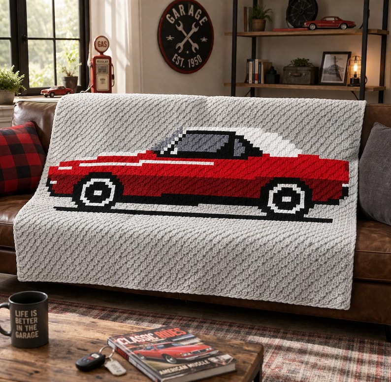 May include: A gray crocheted blanket featuring a red classic car design with a white roof. The blanket is draped over a brown leather couch. A black mug with the text "LIFE IS BETTER IN THE GARAGE" is on a wooden coffee table.