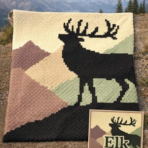 May include: A handmade crochet blanket featuring a black elk silhouette against a backdrop of geometric mountains in shades of brown, green, and cream. The blanket is displayed outdoors with a pattern card that reads "Elk C2C Crochet Blanket Pattern."