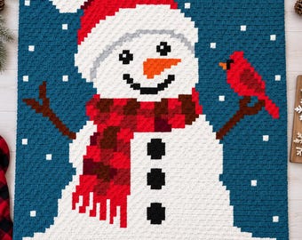 May include: A rectangular crochet blanket featuring a pixelated snowman design against a dark blue background with white snowflakes. The snowman wears a red Santa hat and a red plaid scarf, with three black buttons and a bright orange carrot nose.