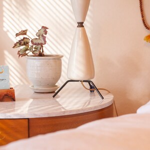 May include: A white ceramic lamp with a black tripod base sits on a marble-topped nightstand. A small wooden stand holds a card with the text "moonyugars" and a gold crescent moon earring. A plant in a white ceramic pot is on the nightstand.