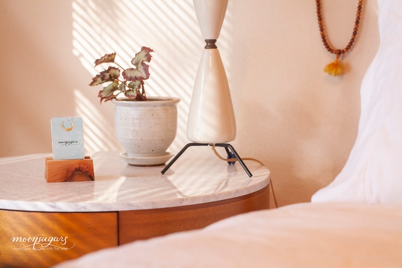 May include: A white ceramic lamp with a black tripod base sits on a marble-topped nightstand. A small wooden stand holds a card with the text "moonyugars" and a gold crescent moon earring. A plant in a white ceramic pot is on the nightstand.